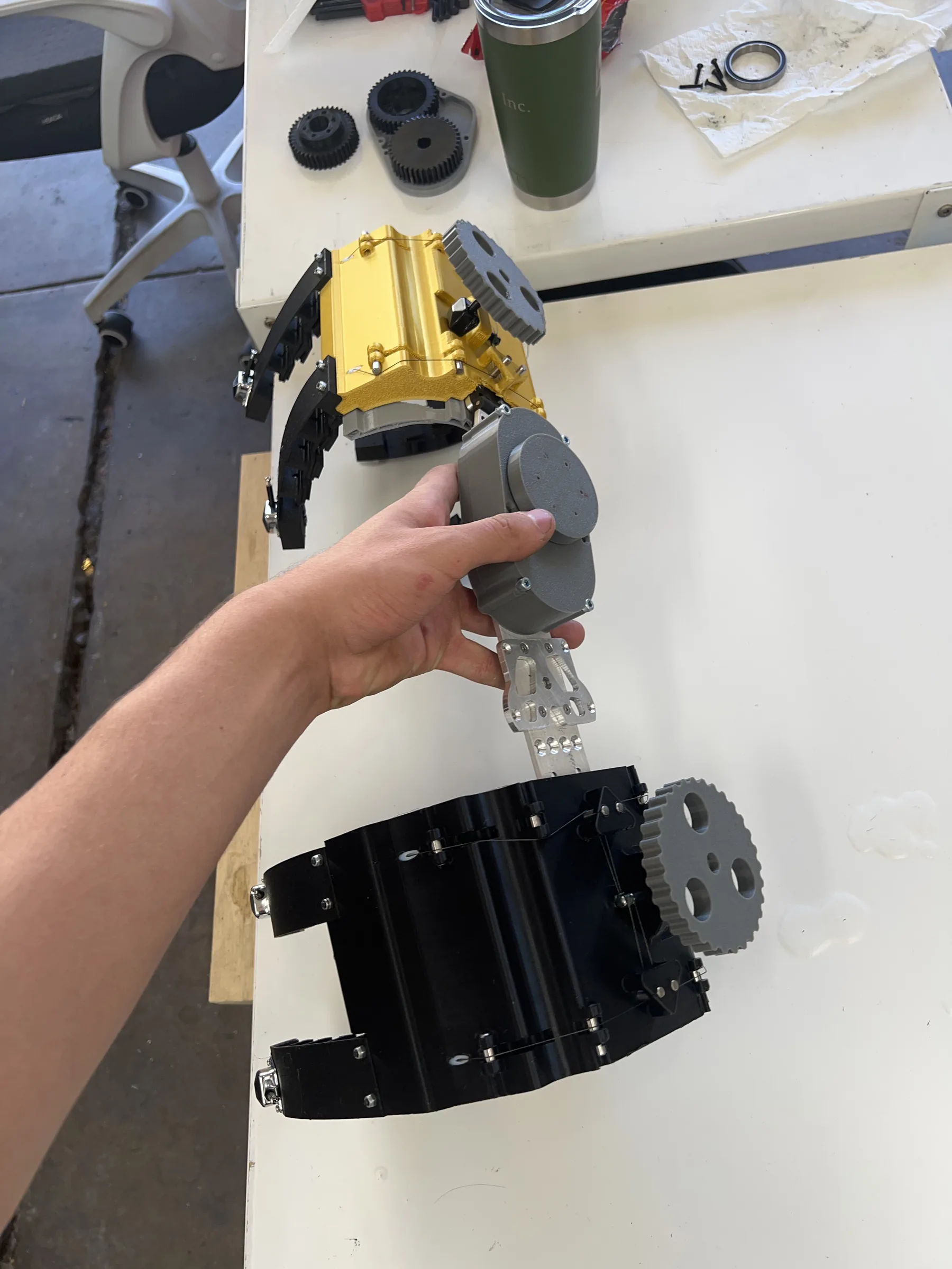 Hand holding the finished leg brace prototype: a yellow and black 3D-printed body with curved black finger straps, gray tuning-peg dials and a machined aluminium link plate