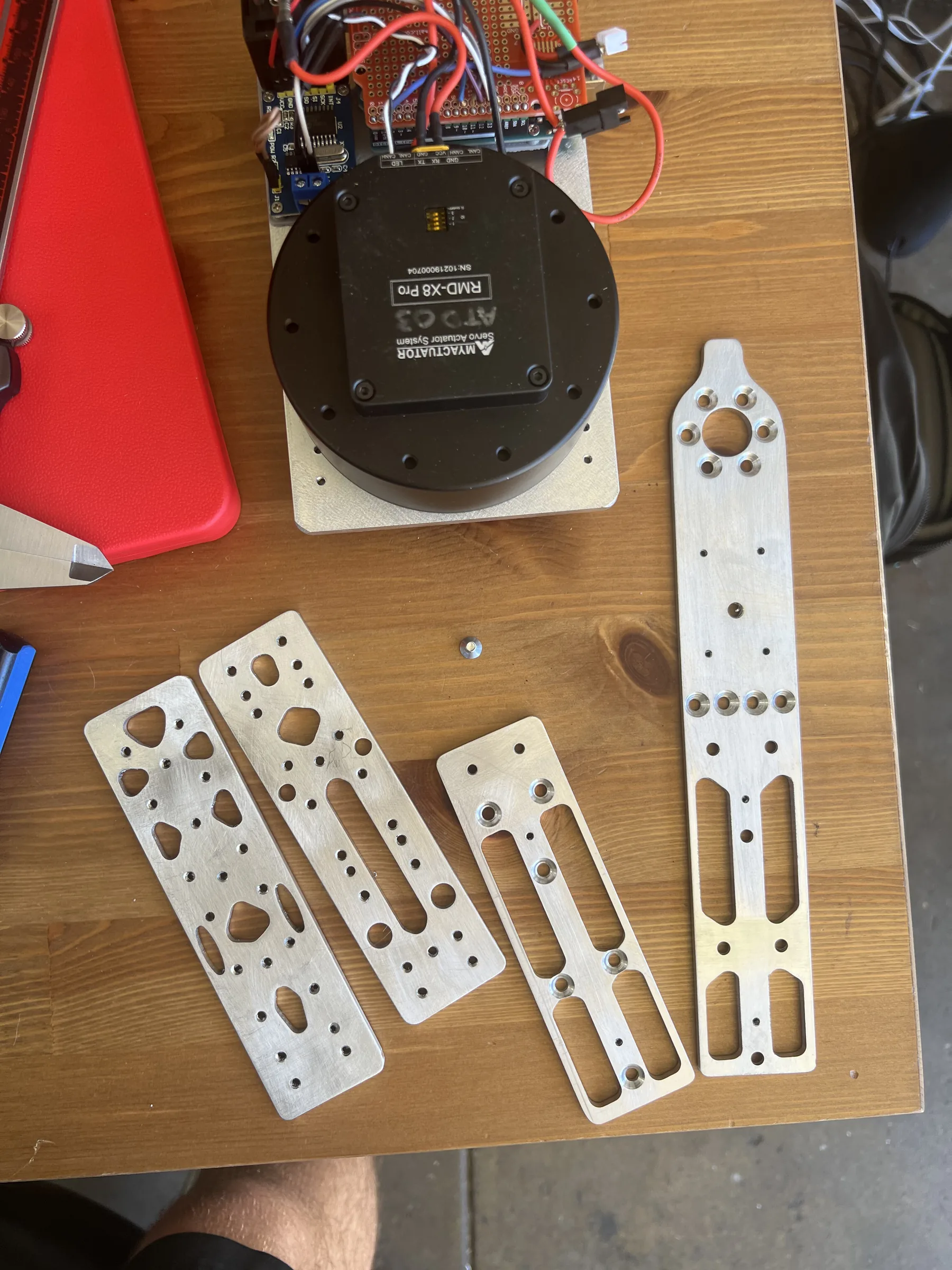 Four CNC-machined aluminium plates with weight-saving cutouts laid on a wooden bench next to an RMD-X8 Pro servo actuator and its wiring