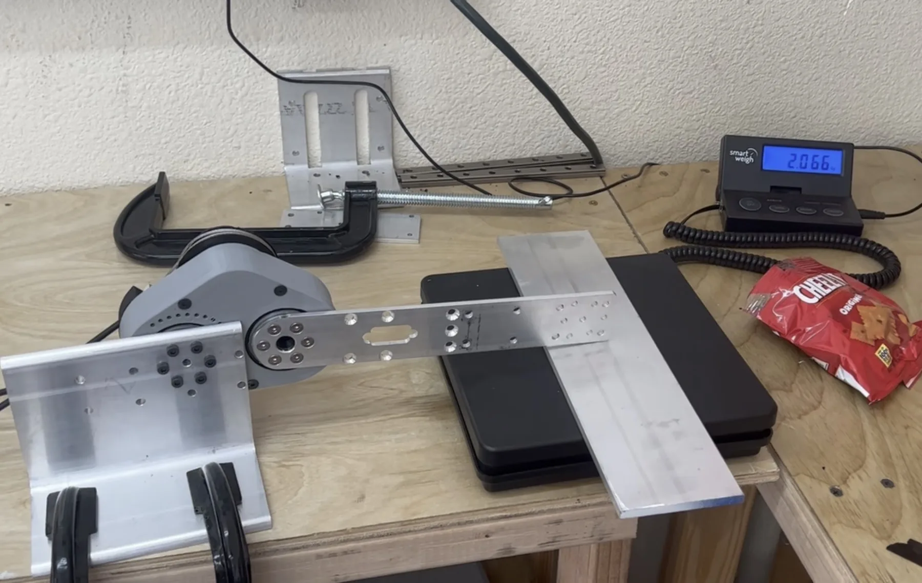 Force test rig on a plywood bench: a brace joint clamped to an aluminium bracket presses a machined arm onto a digital scale reading 2.066 kg