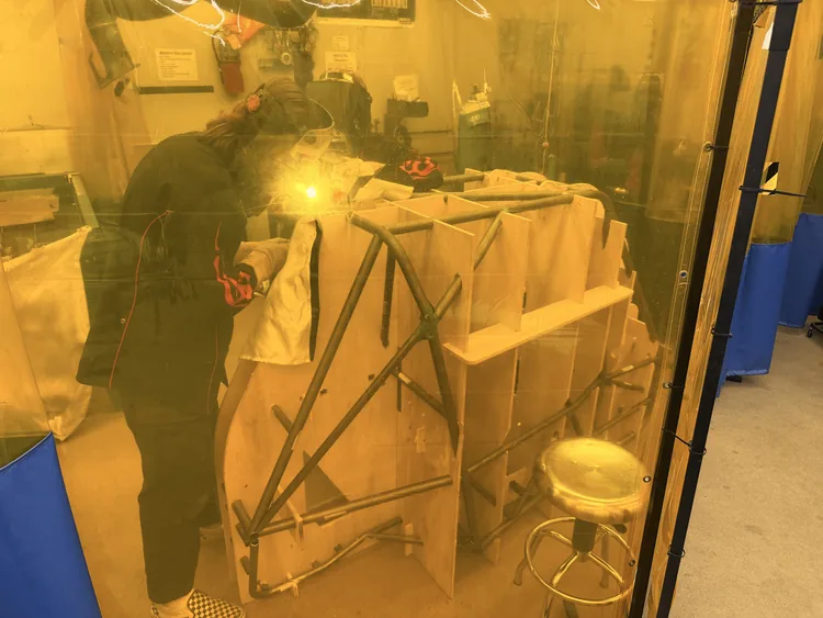 A team member in a welding helmet TIG welding the chassis frame inside a curtained welding booth, arc glowing