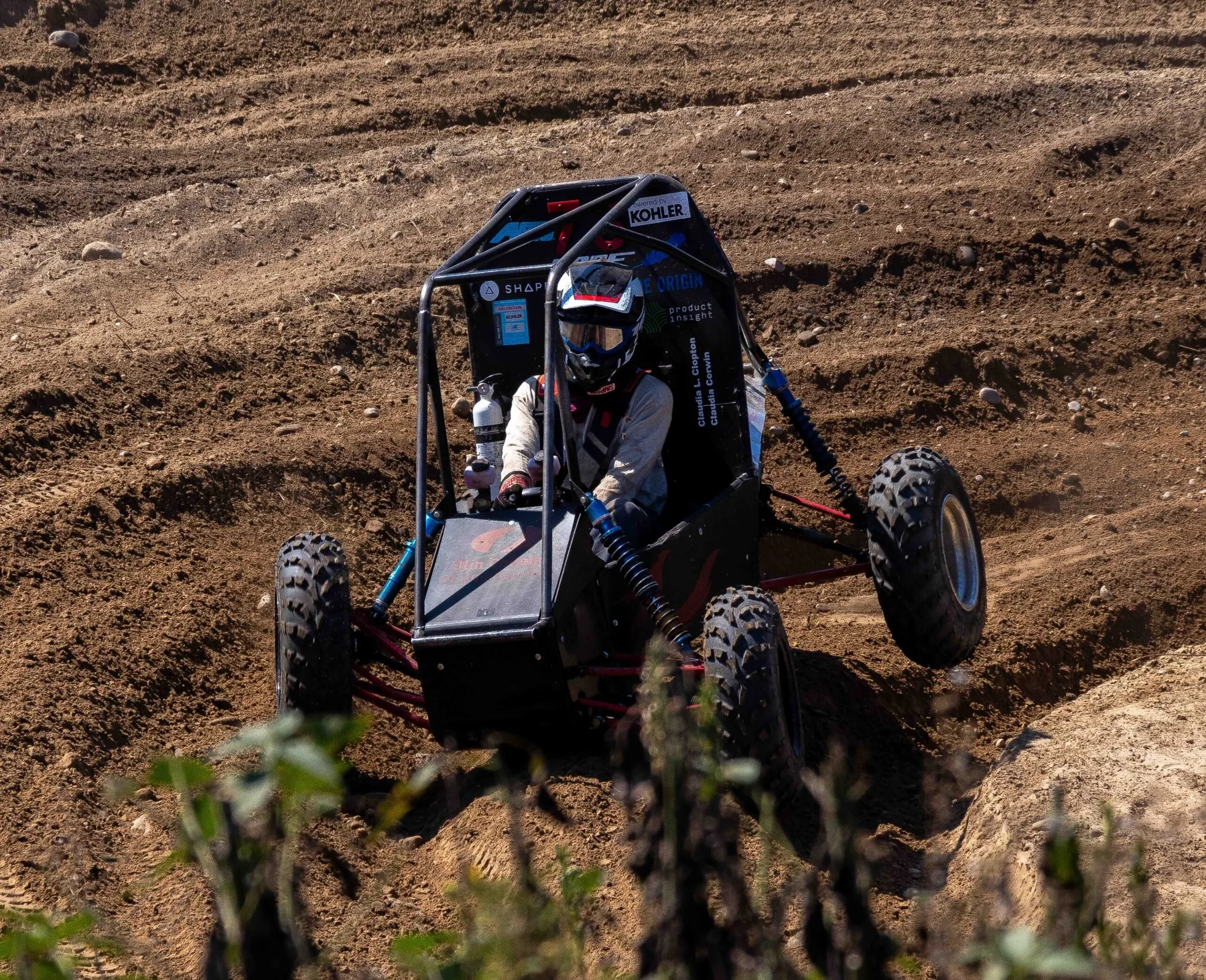 The Olin Baja car with a helmeted driver cresting a dirt mound on a rough off-road course