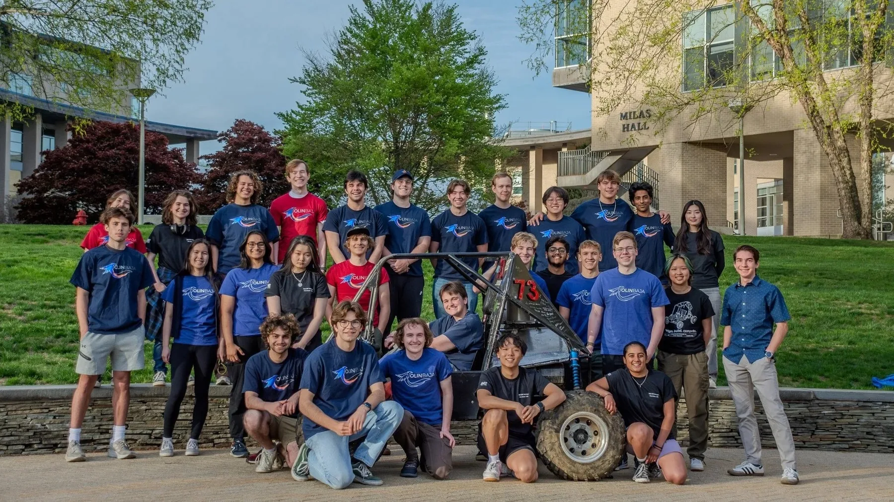 The Olin Baja SAE team in matching blue and red shirts posing outdoors around the off-road car, with a wheel held in front