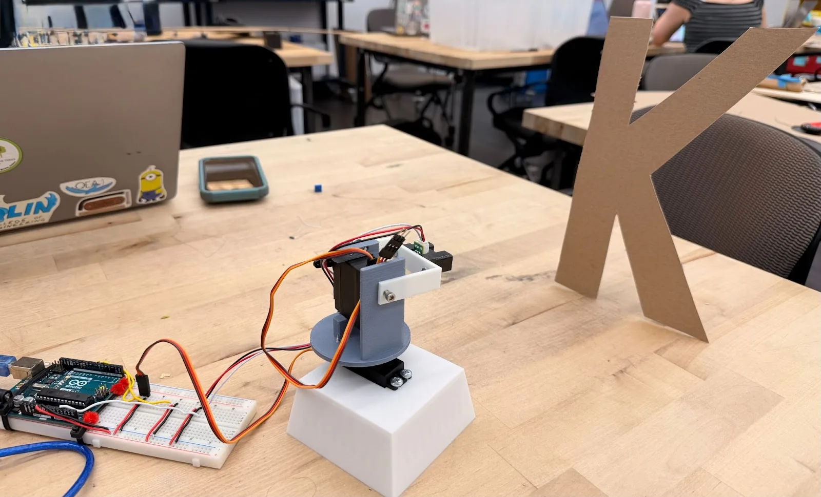 The scanner on a workshop table pointed at a large cardboard letter K standing upright, with the Arduino and breadboard beside it and a laptop in the background