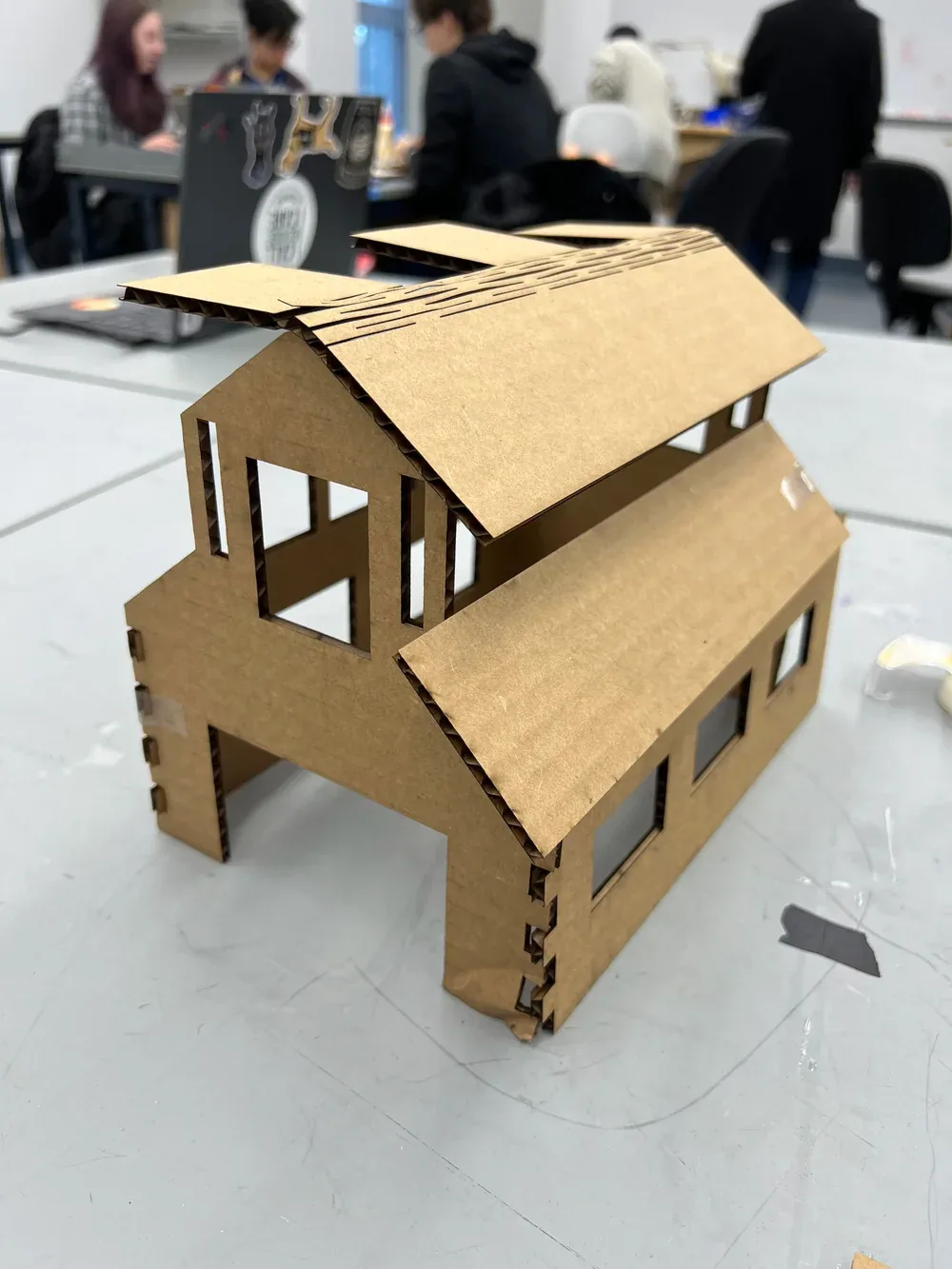 Cardboard prototype of the barn with a slatted roof and cut-out windows on a workshop table