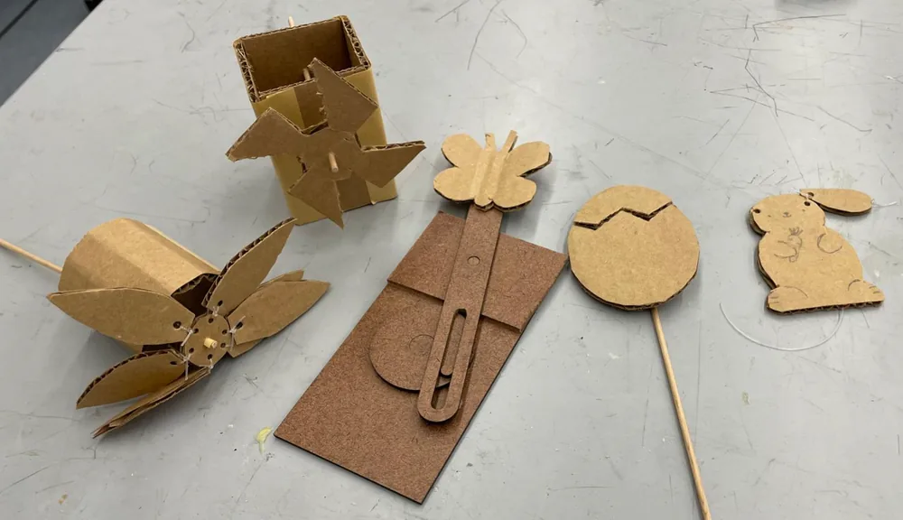 Cardboard prototypes of the windmill, butterfly, flower linkage, cracked egg and bunny laid out on a table
