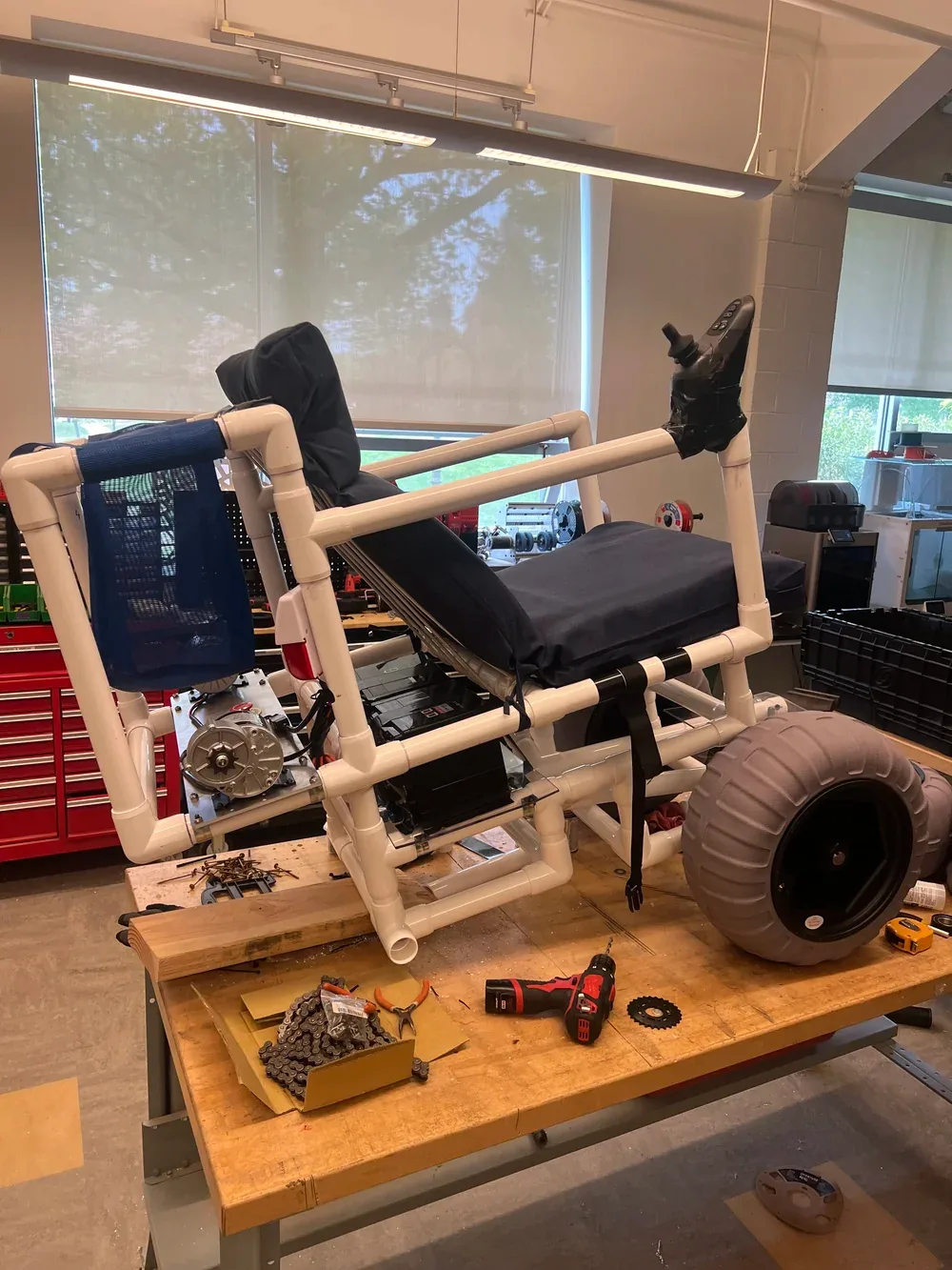 The wheelchair mid-build on a workshop bench: the PVC frame with seat cushion and joystick fitted, a silver electric motor and battery box mounted under the seat, one balloon tire attached, and a drill, chain and sprocket on the bench