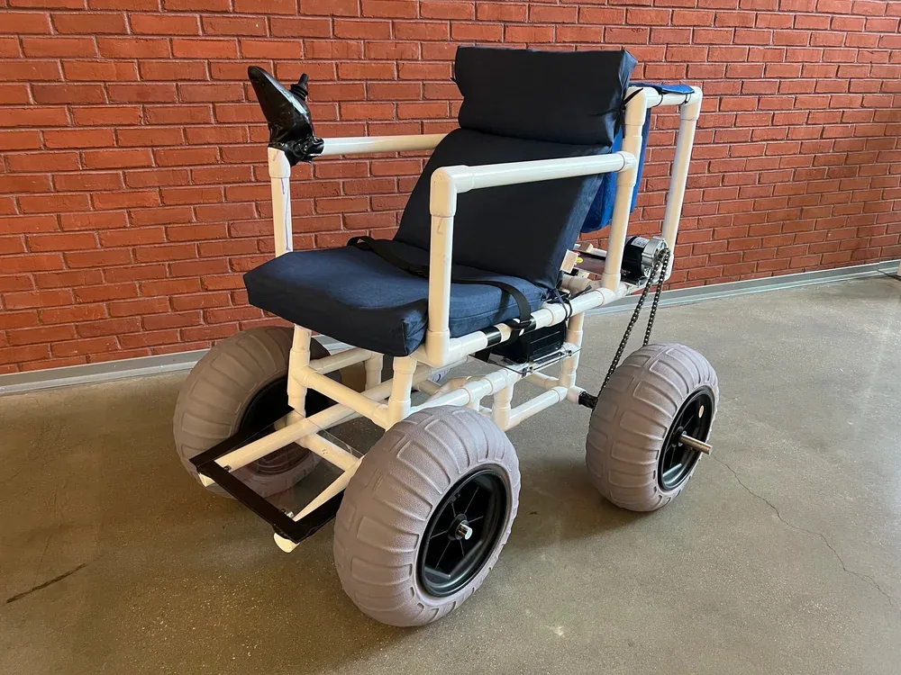 The finished motorized beach wheelchair in front of a brick wall: a white PVC frame with a navy cushioned seat and back, a joystick on the armrest, four wide gray balloon beach tires and a chain drive to the rear axle
