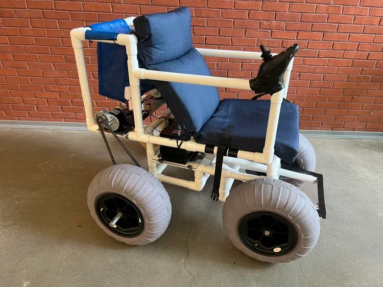 Side view of the finished motorized beach wheelchair against a brick wall, showing the chain drive from the motor to the rear balloon tire, the battery box under the seat and the joystick on the right armrest